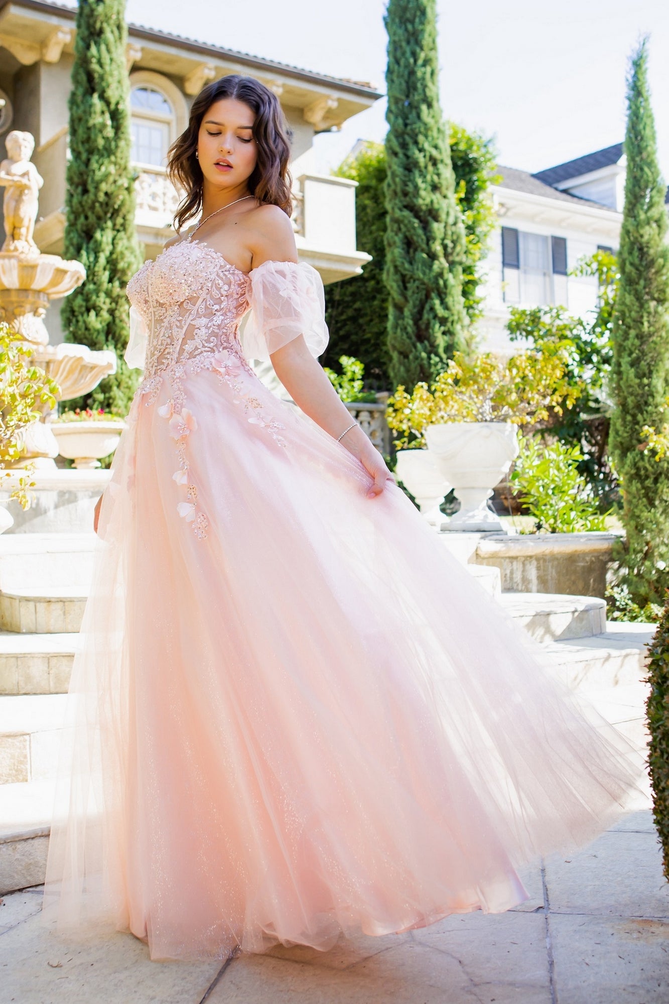 A woman in the Puff-Sleeve Long Sweetheart Prom Ball Gown 8042J—light pink with off-the-shoulder tulle, puff sleeves, and beaded floral details—stands on stone steps outdoors amid greenery and elegant architecture.